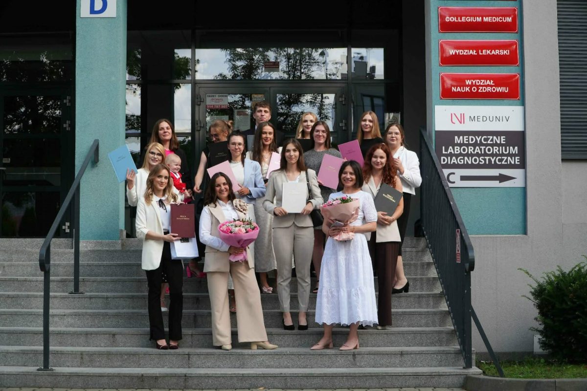 Zdjęcie przedstawia grupę uczestników pozujących do wspólnego zdjęcia przed budynkiem uczelni lub instytucji. Część osób trzyma dyplomy, certyfikaty oraz kwiaty, podkreślając uroczysty charakter wydarzenia. W tle widoczne jest wejście do budynku oraz tablice informacyjne. Fotografia upamiętnia wspólne spotkanie lub zakończenie ważnego etapu.