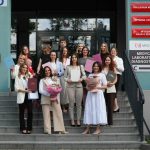 Zdjęcie przedstawia grupę uczestników pozujących do wspólnego zdjęcia przed budynkiem uczelni lub instytucji. Część osób trzyma dyplomy, certyfikaty oraz kwiaty, podkreślając uroczysty charakter wydarzenia. W tle widoczne jest wejście do budynku oraz tablice informacyjne. Fotografia upamiętnia wspólne spotkanie lub zakończenie ważnego etapu.