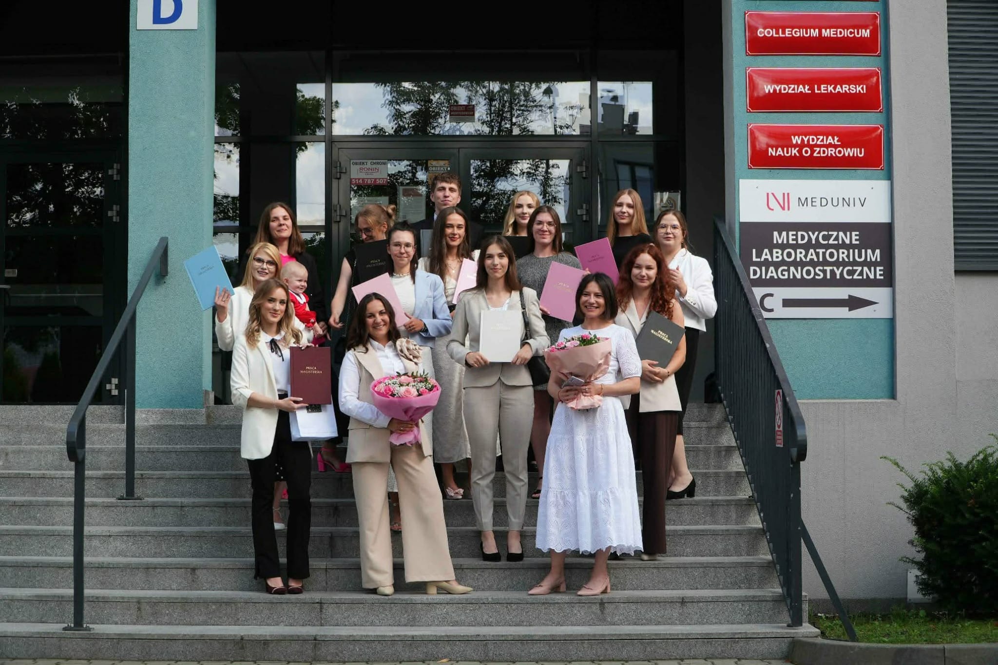 Zdjęcie przedstawia grupę uczestników pozujących do wspólnego zdjęcia przed budynkiem uczelni lub instytucji. Część osób trzyma dyplomy, certyfikaty oraz kwiaty, podkreślając uroczysty charakter wydarzenia. W tle widoczne jest wejście do budynku oraz tablice informacyjne. Fotografia upamiętnia wspólne spotkanie lub zakończenie ważnego etapu.