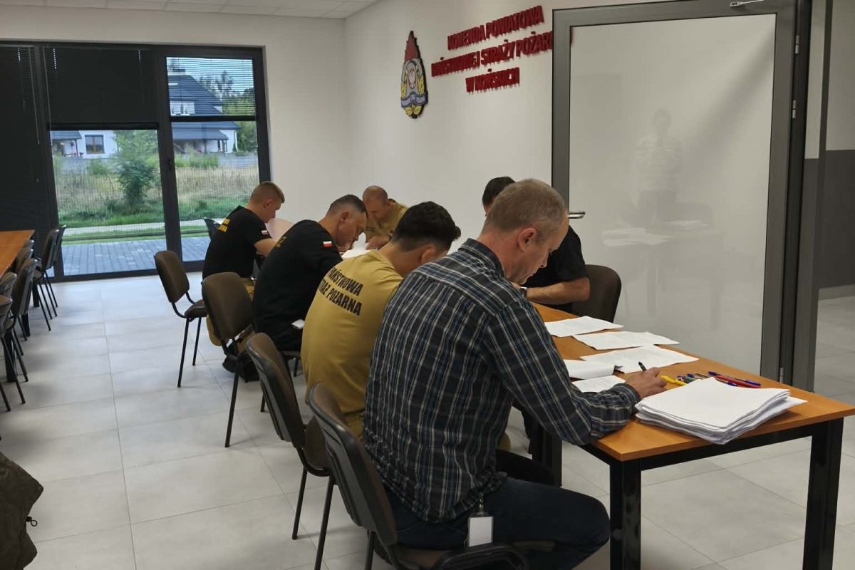 Zdjęcie przedstawia jasną salę dydaktyczną, w której uczestnicy pracują przy drewnianym stole z dokumentami i materiałami szkoleniowymi. Na ścianie widoczne są oznaczenia związane ze służbami ratowniczymi. Duże okna zapewniają dostęp do naturalnego światła, a w tle widoczne jest wyposażenie techniczne sali.