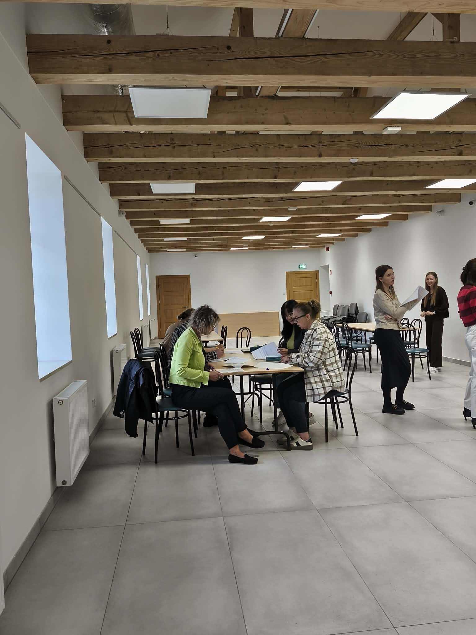 Zdjęcie przedstawia jasną, przestronną salę dydaktyczną z drewnianymi stołami i krzesłami. Uczestnicy siedzą przy stołach, pracując z dokumentami i rozmawiając. Duże okna oraz oświetlenie sufitowe zapewniają dobre doświetlenie pomieszczenia.
