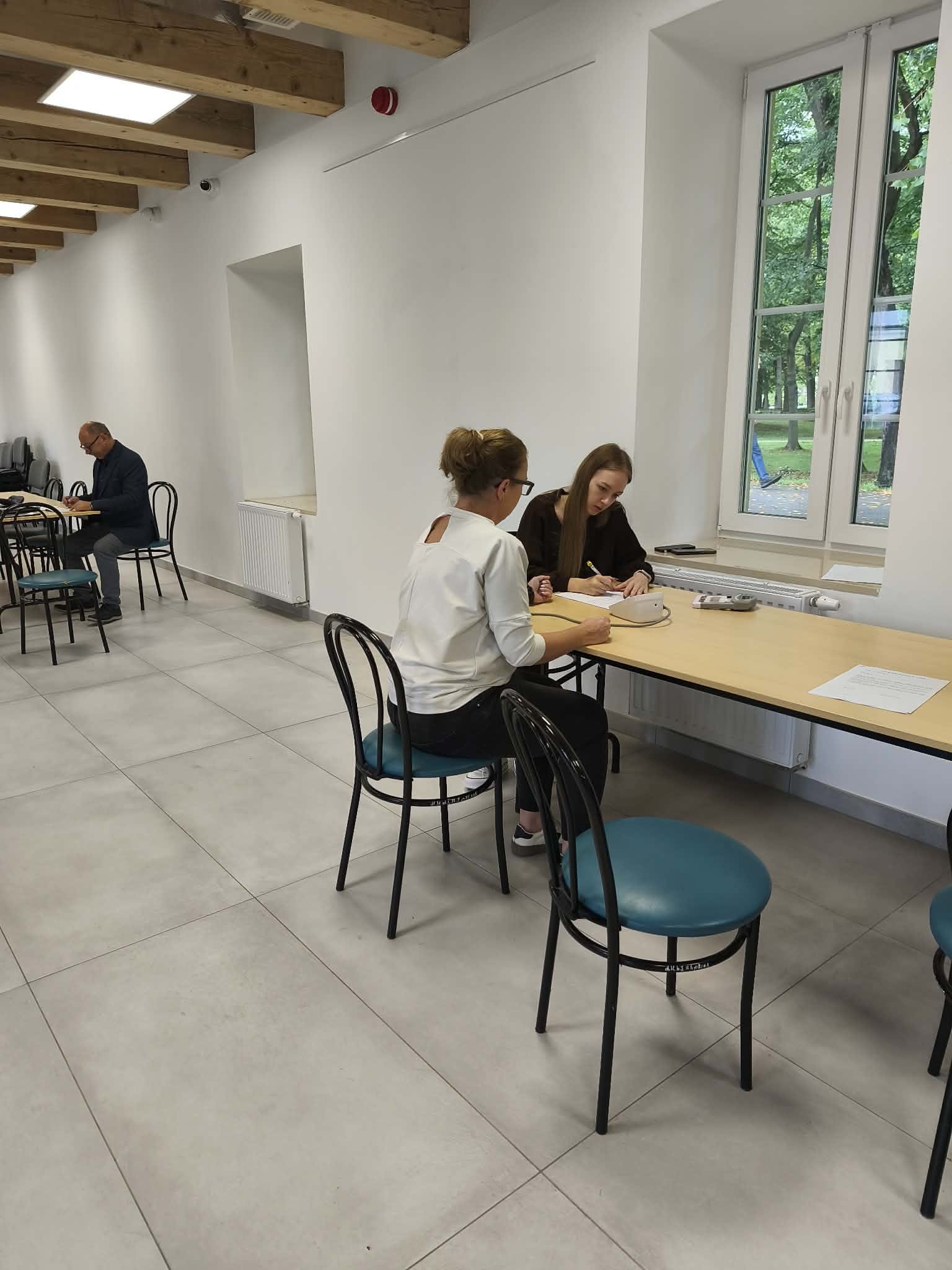 Zdjęcie przedstawia jasną, nowoczesną salę wyposażoną w drewniane stoły i krzesła. W pomieszczeniu widoczni są uczestnicy pracujący przy stołach z wykorzystaniem dokumentów i materiałów dydaktycznych. Duże okna zapewniają dostęp do naturalnego światła, a za nimi widoczna jest zieleń.