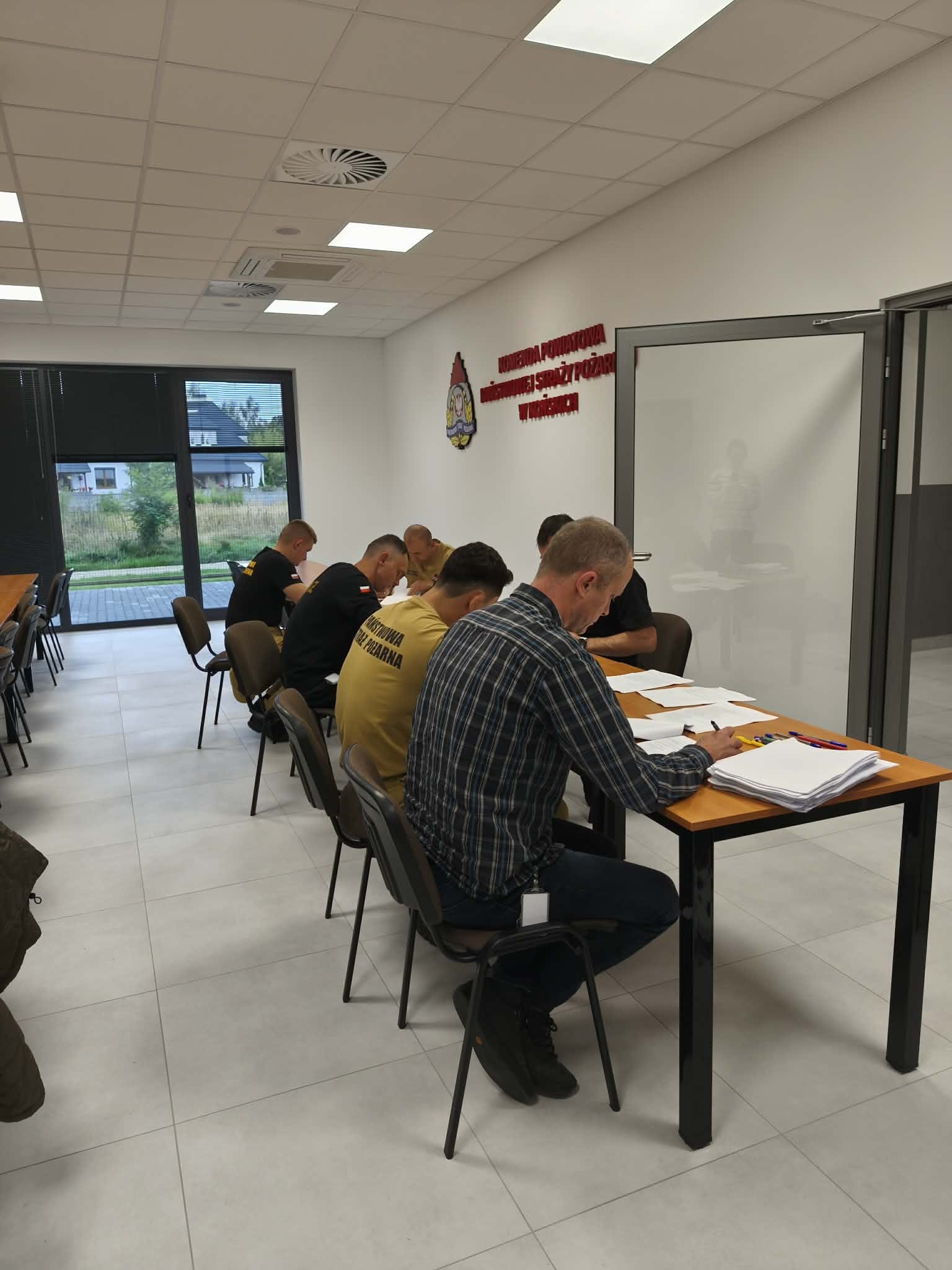Zdjęcie przedstawia jasną salę dydaktyczną, w której uczestnicy pracują przy drewnianym stole z dokumentami i materiałami szkoleniowymi. Na ścianie widoczne są oznaczenia związane ze służbami ratowniczymi. Duże okna zapewniają dostęp do naturalnego światła, a w tle widoczne jest wyposażenie techniczne sali.