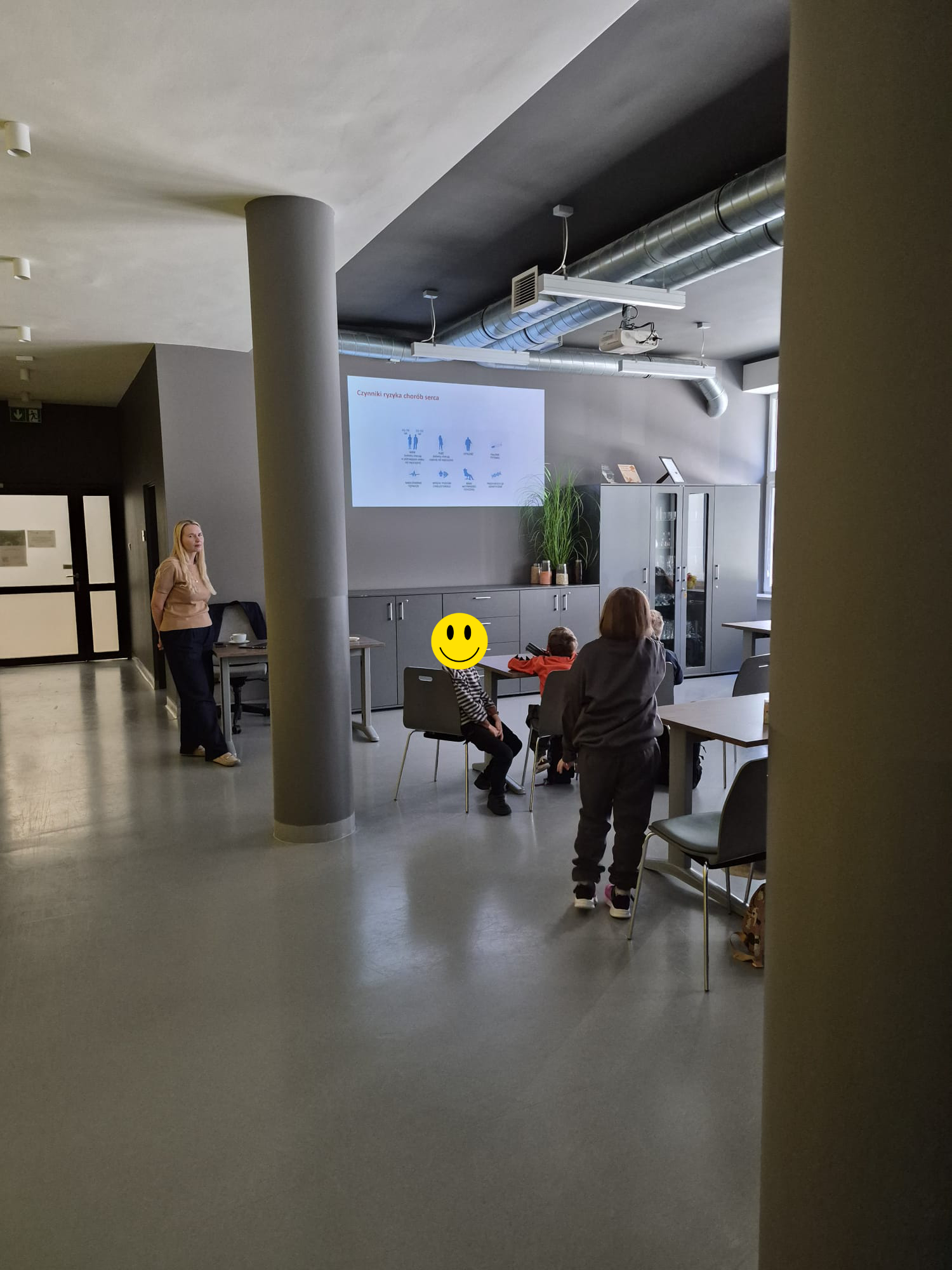 Zdjęcie przedstawia nowoczesną salę dydaktyczną, w kt&oacute;rej uczestnicy oglądają prezentację wyświetlaną na tylnej ścianie. Na ekranie widoczny jest polski nagł&oacute;wek oraz niebieskie ikony związane ze zdrowiem. W sali znajdują się stoły, krzesła, szafki, rośliny, okna oraz instalacja wentylacyjna.