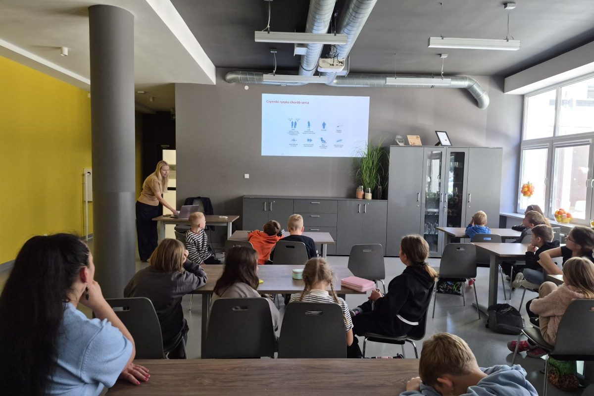 Zdjęcie przedstawia prezentację edukacyjną prowadzoną w sali dydaktycznej, z wykorzystaniem laptopa i projektora. Na ekranie wyświetlana jest prezentacja z czerwonym nagłówkiem oraz ikonami związanymi ze zdrowiem. W sali znajdują się stoły, krzesła, szafki, rośliny oraz wyposażenie techniczne.