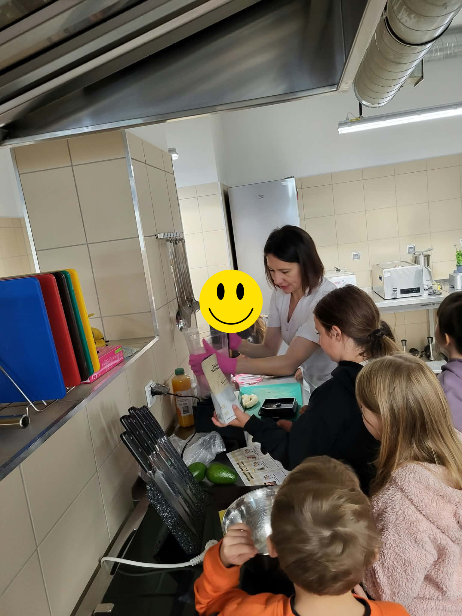 Zdjęcie przedstawia zajęcia kulinarne w kuchni dydaktycznej, podczas kt&oacute;rych uczestnicy przygotowują żywność przy stanowisku roboczym. Na blacie znajdują się miska, limonki, przybory kuchenne, deski do krojenia oraz opakowania produkt&oacute;w. W tle widoczne są kolorowe deski, lod&oacute;wka, zlew, sprzęt kuchenny i instalacja wentylacyjna.