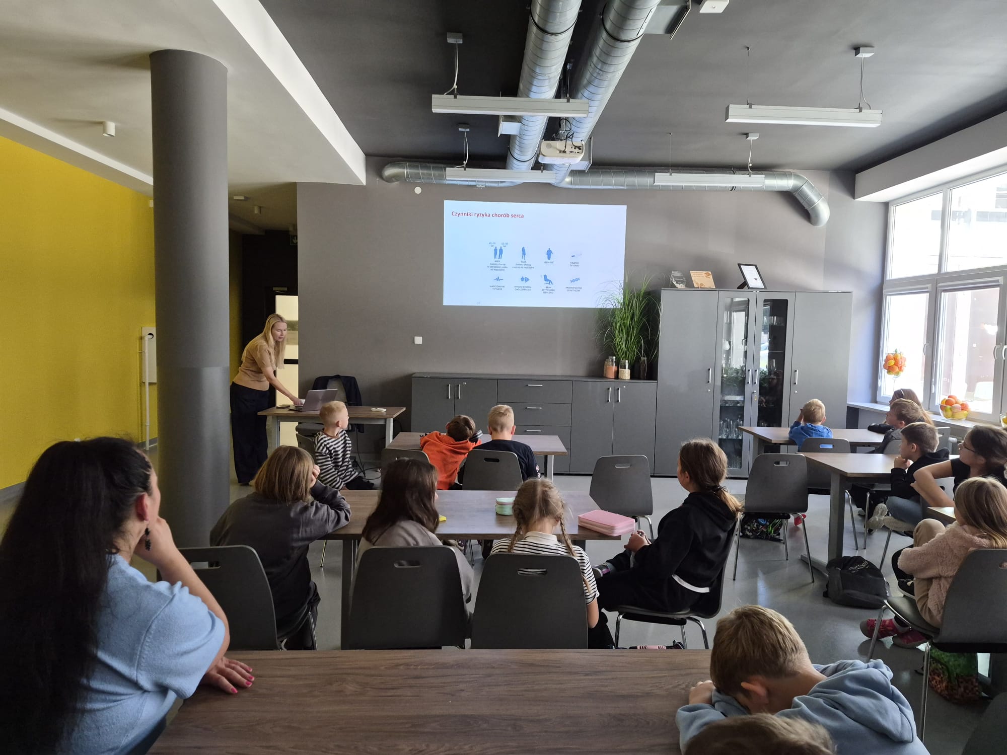 Zdjęcie przedstawia prezentację edukacyjną prowadzoną w sali dydaktycznej, z wykorzystaniem laptopa i projektora. Na ekranie wyświetlana jest prezentacja z czerwonym nagł&oacute;wkiem oraz ikonami związanymi ze zdrowiem. W sali znajdują się stoły, krzesła, szafki, rośliny oraz wyposażenie techniczne.