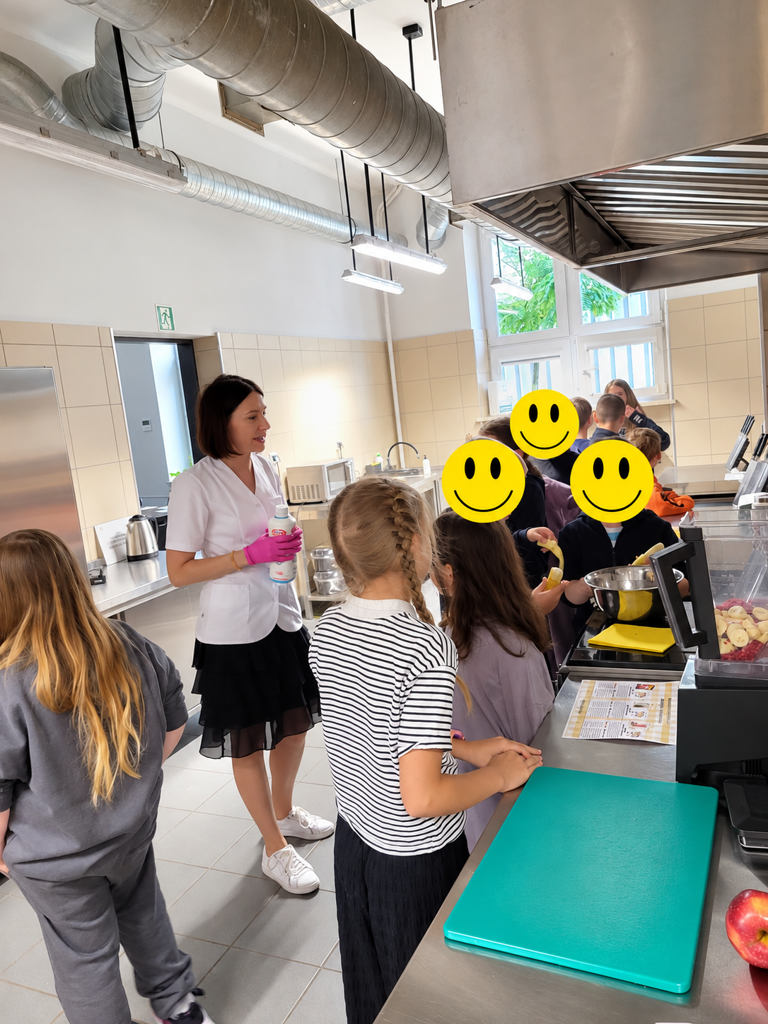 Zdjęcie przedstawia zajęcia kulinarne w profesjonalnej kuchni, podczas kt&oacute;rych uczestnicy przygotowują żywność przy stalowych stanowiskach roboczych. Na blatach znajdują się miski, deski do krojenia, przybory kuchenne oraz przygotowane owoce. W tle widoczne są urządzenia kuchenne, okapy wentylacyjne, oświetlenie i płytki ścienne.