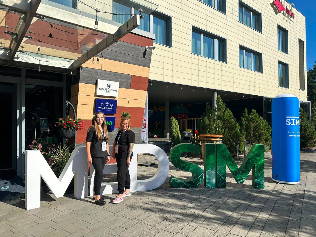 Dwie osoby stoją przed dużą instalacją z literami &bdquo;MDSM&rdquo; ustawioną przy wejściu do nowoczesnego budynku hotelowego. W tle widoczne są tablice z napisami &bdquo;Grape Town Hotel&rdquo; i &bdquo;Winogrono Restauracja&rdquo;, szklane zadaszenie oraz ozdobne rośliny. Po prawej stronie znajduje się niebieski słup reklamowy z napisem &bdquo;SIM&rdquo;. Zdjęcie wykonano w słoneczny dzień na zewnątrz budynku.
