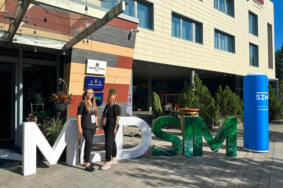Dwie osoby stoją przed dużą instalacją z literami „MDSM” ustawioną przy wejściu do nowoczesnego budynku hotelowego. W tle widoczne są tablice z napisami „Grape Town Hotel” i „Winogrono Restauracja”, szklane zadaszenie oraz ozdobne rośliny. Po prawej stronie znajduje się niebieski słup reklamowy z napisem „SIM”. Zdjęcie wykonano w słoneczny dzień na zewnątrz budynku.