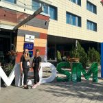 Dwie osoby stoją przed dużą instalacją z literami „MDSM” ustawioną przy wejściu do nowoczesnego budynku hotelowego. W tle widoczne są tablice z napisami „Grape Town Hotel” i „Winogrono Restauracja”, szklane zadaszenie oraz ozdobne rośliny. Po prawej stronie znajduje się niebieski słup reklamowy z napisem „SIM”. Zdjęcie wykonano w słoneczny dzień na zewnątrz budynku.