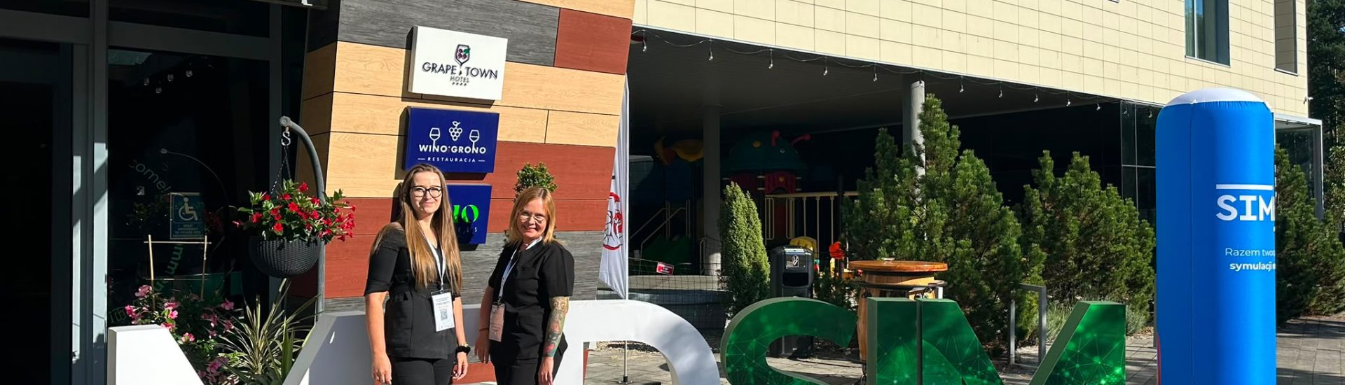 Dwie osoby stoją przed dużą instalacją z literami „MDSM” ustawioną przy wejściu do nowoczesnego budynku hotelowego. W tle widoczne są tablice z napisami „Grape Town Hotel” i „Winogrono Restauracja”, szklane zadaszenie oraz ozdobne rośliny. Po prawej stronie znajduje się niebieski słup reklamowy z napisem „SIM”. Zdjęcie wykonano w słoneczny dzień na zewnątrz budynku.