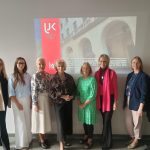 Zdjęcie przedstawia grupę uczestniczek pozujących do wspólnego zdjęcia na tle prezentacji przedstawiającej budynek uczelni. W tle widoczne jest logo i nazwa uniwersytetu. Fotografia została wykonana podczas spotkania lub wydarzenia akademickiego.