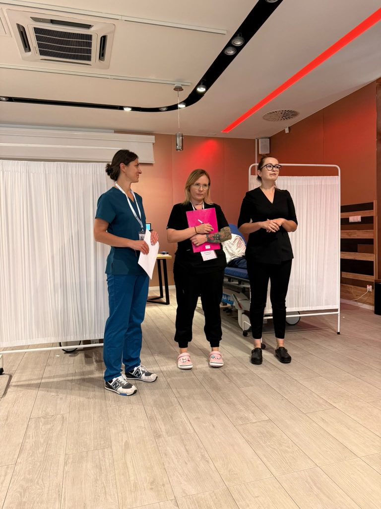 Trzy osoby stoją w sali szkoleniowej zaaranżowanej na stanowisko medyczne. W tle znajdują się parawany oraz ł&oacute;żko szpitalne. Jedna osoba trzyma dokumenty, druga r&oacute;żową teczkę, a trzecia przemawia do uczestnik&oacute;w. Pomieszczenie jest nowoczesne, z jasną podłogą i oświetleniem sufitowym.