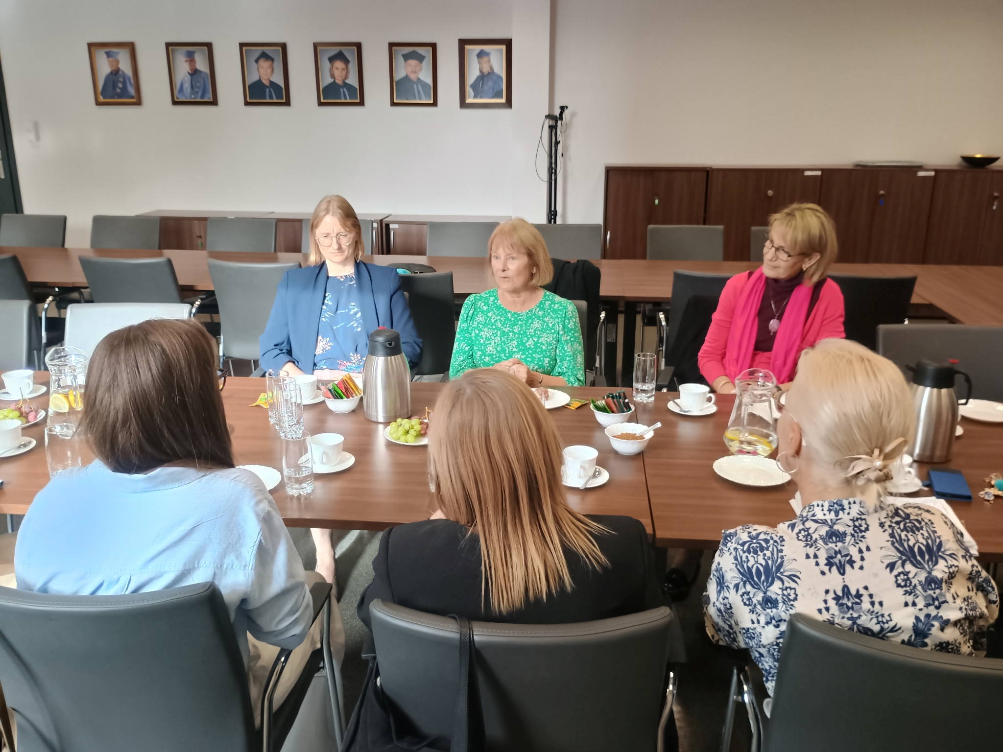 Zdjęcie przedstawia spotkanie uczestnik&oacute;w przy długim stole konferencyjnym. Na stole widoczne są materiały, napoje, owoce oraz przybory wykorzystywane podczas spotkania. W tle znajdują się szafy, oprawione fotografie oraz sprzęt do rejestracji wydarzenia.