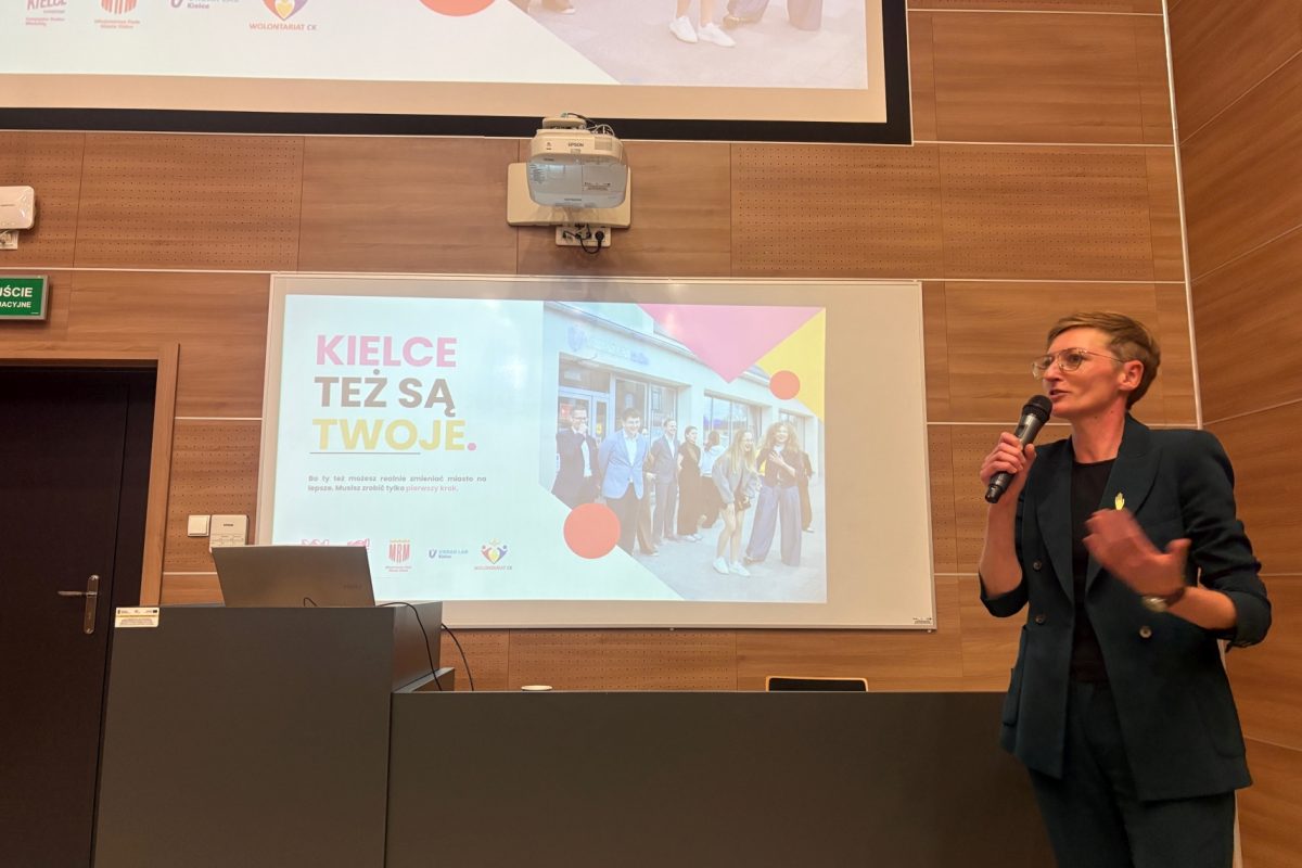 Zdjęcie przedstawia wystąpienie w sali wykładowej, podczas którego prelegentka przemawia do uczestników z wykorzystaniem mikrofonu. Na ekranie wyświetlana jest kolorowa prezentacja w języku polskim z fotografią grupową i geometrycznymi elementami graficznymi. W tle widoczne są drewniane panele ścienne, mównica z laptopem oraz projektor sufitowy.