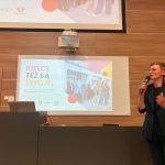 Zdjęcie przedstawia wystąpienie w sali wykładowej, podczas którego prelegentka przemawia do uczestników z wykorzystaniem mikrofonu. Na ekranie wyświetlana jest kolorowa prezentacja w języku polskim z fotografią grupową i geometrycznymi elementami graficznymi. W tle widoczne są drewniane panele ścienne, mównica z laptopem oraz projektor sufitowy.