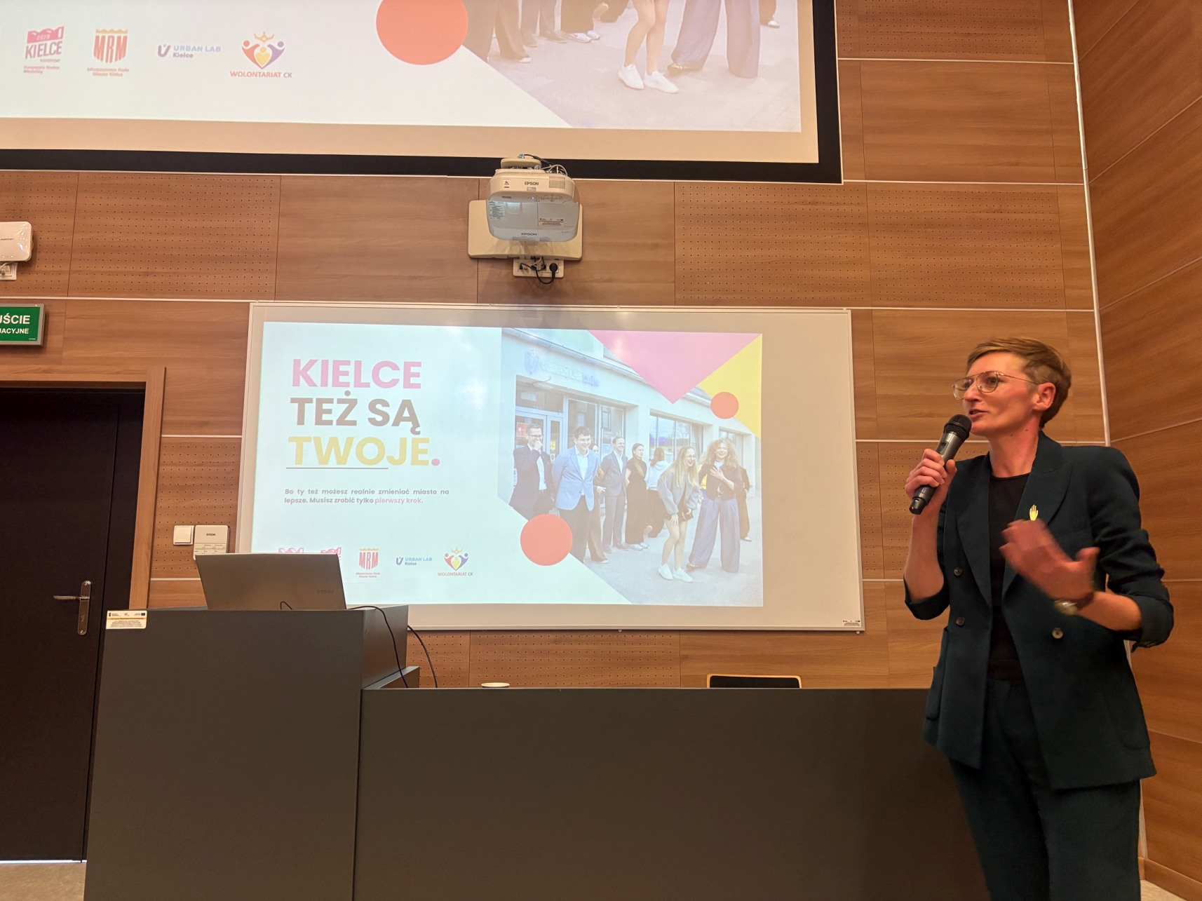 Zdjęcie przedstawia wystąpienie w sali wykładowej, podczas którego prelegentka przemawia do uczestników z wykorzystaniem mikrofonu. Na ekranie wyświetlana jest kolorowa prezentacja w języku polskim z fotografią grupową i geometrycznymi elementami graficznymi. W tle widoczne są drewniane panele ścienne, mównica z laptopem oraz projektor sufitowy.